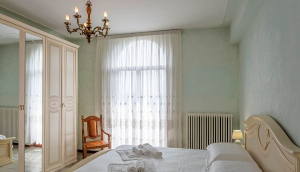 A hotel room with a bed, a chair, a wardrobe, a radiator, and a chandelier.