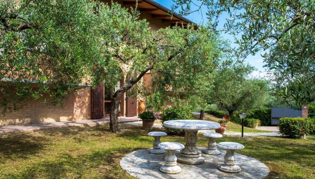 Une cour avec une table et des chaises en pierre, entourée d’arbres et d’un bâtiment.