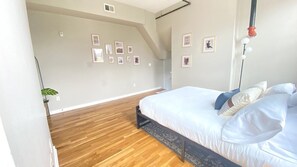 A bedroom with a wooden floor, a bed with white bedding, and a wall with framed pictures.