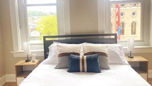 Habitación de hotel con una cama grande, dos mesitas de noche y un ventanal con vistas a un edificio y árboles.
