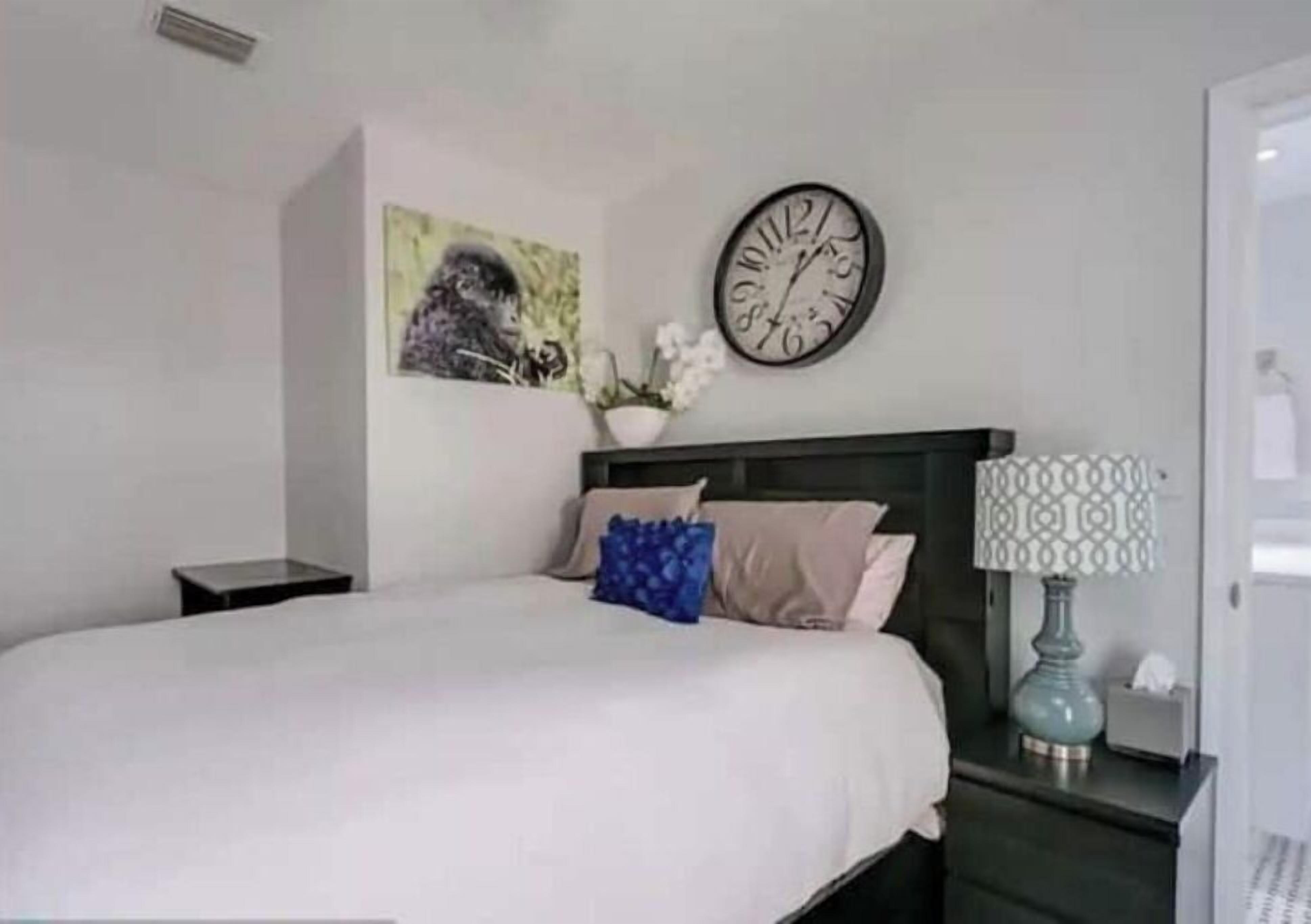 Een netjes opgemaakt bed met een witte deken en twee kussens, een wandklok, een klein nachtkastje met een lamp en een ingelijst schilderij aan de muur.