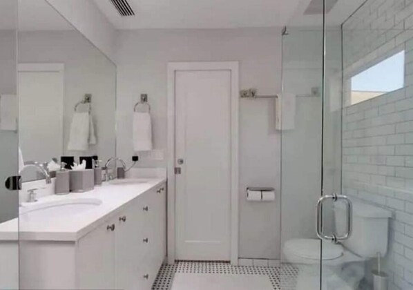 Une salle de bain moderne dotée d’une douche à parois de verre, d’un lavabo blanc avec un grand miroir et de toilettes séparées par une cloison en verre.