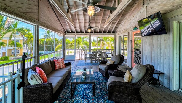 A covered outdoor seating area with a ceiling fan, a TV mounted on the wall, and a view of the outdoors. - Coral Cove: Walk to the Beach! Heated pool! Prime North End! (Anna Maria)