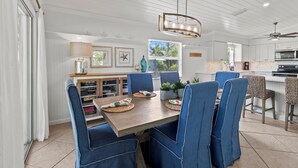 A dining area with a wooden table, blue upholstered chairs, and a hanging light fixture. - Coral Cove: Walk to the Beach! Heated pool! Prime North End! (Anna Maria)