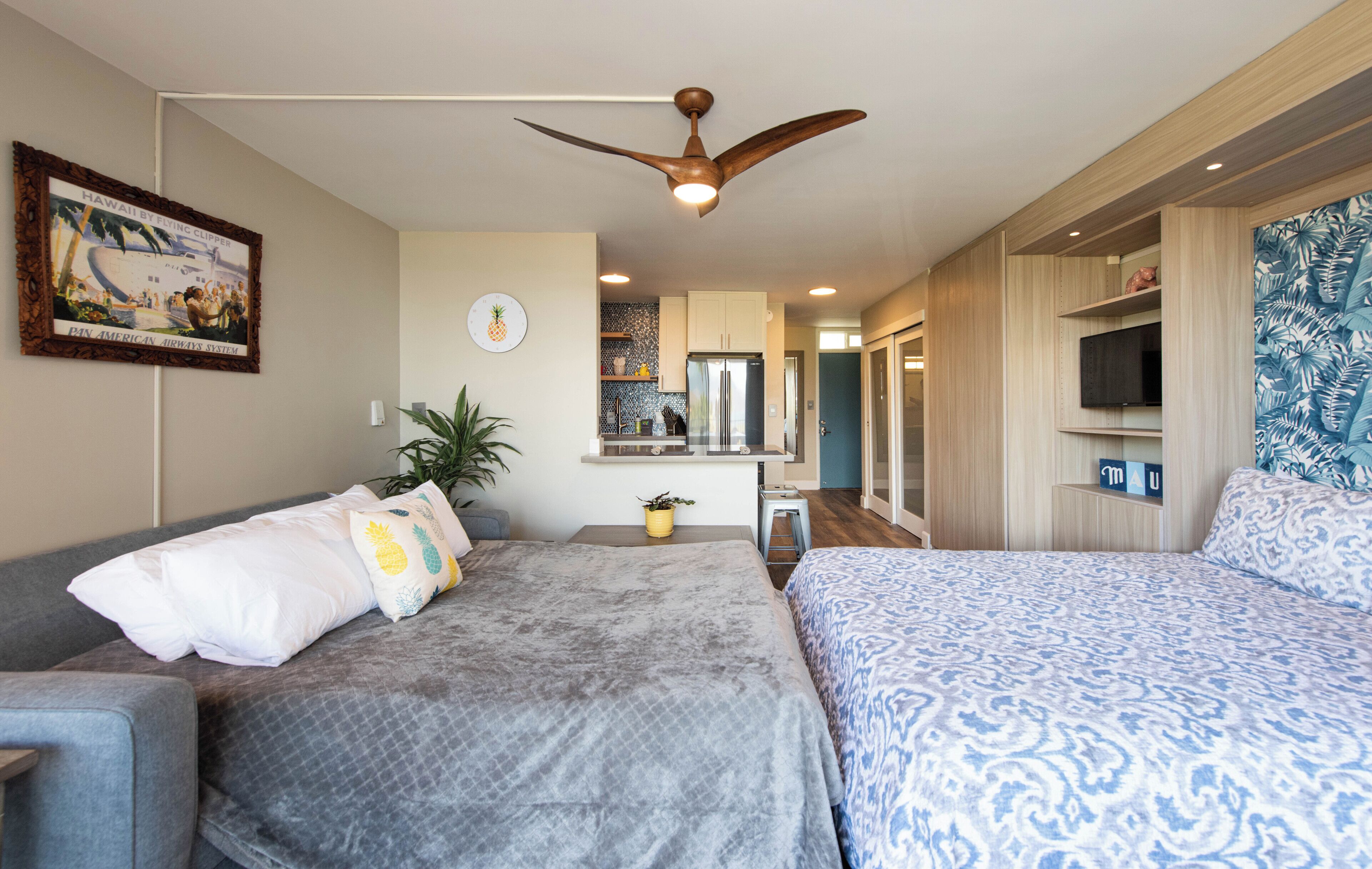 A hotel room with two beds, a ceiling fan, a kitchen area, and a TV on the wall.