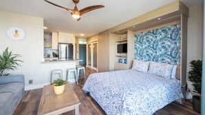 Une chambre moderne avec un grand lit, un bureau en bois, un ventilateur de plafond et un coin cuisine à l’arrière-plan.