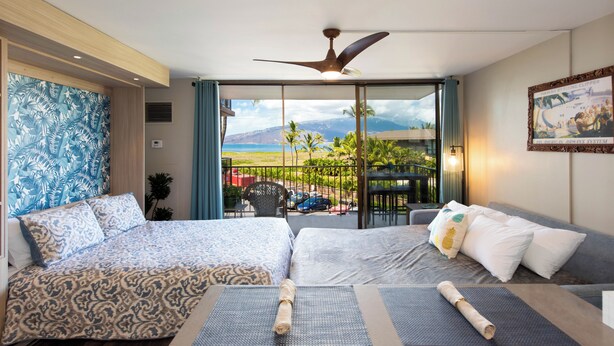Une chambre d’hôtel avec un lit, un canapé, un balcon donnant sur la plage et les palmiers, et un ventilateur de plafond.