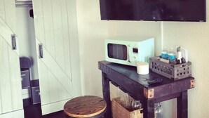 Una habitación con puerta corredera, televisor montado, microondas, un taburete pequeño y una mesa con varios objetos.