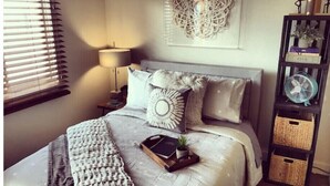 Un dormitorio ordenado con cama, lámpara de noche, adorno en la pared y una pequeña estantería con objetos.