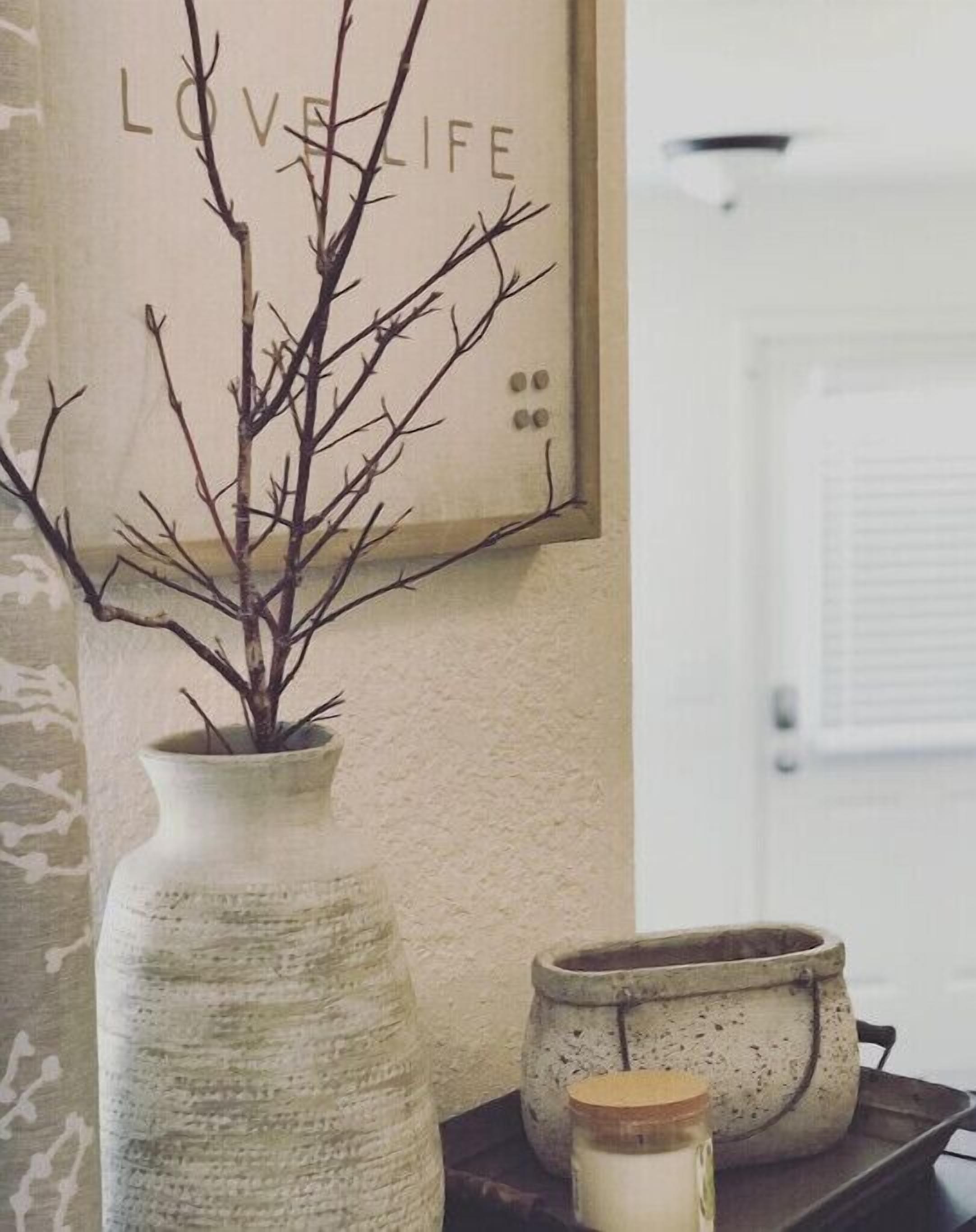 A vase with a branch on a shelf, a framed sign with the words "LOVE LIFE", and a candle in a jar on a tray.