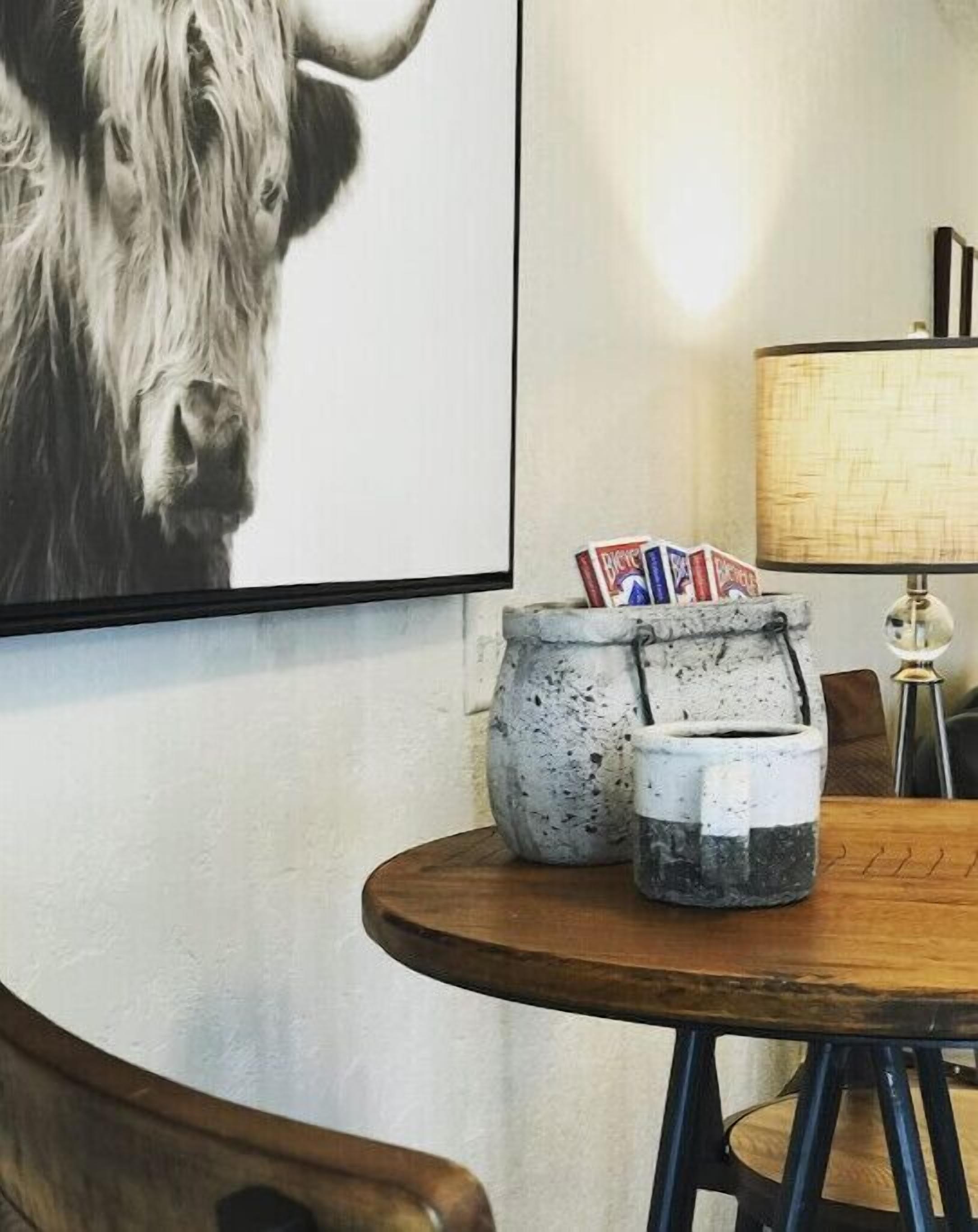 A round wooden table with two ceramic containers holding playing cards, a lamp, and a framed picture of a bison on the wall.