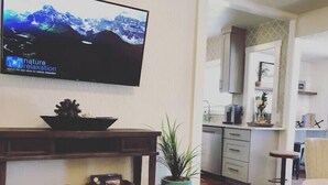 A living room with a television displaying 'nature relaxation', a wooden coffee table with decorative items, a kitchen in the background, and a dining area with a cowhide rug.
