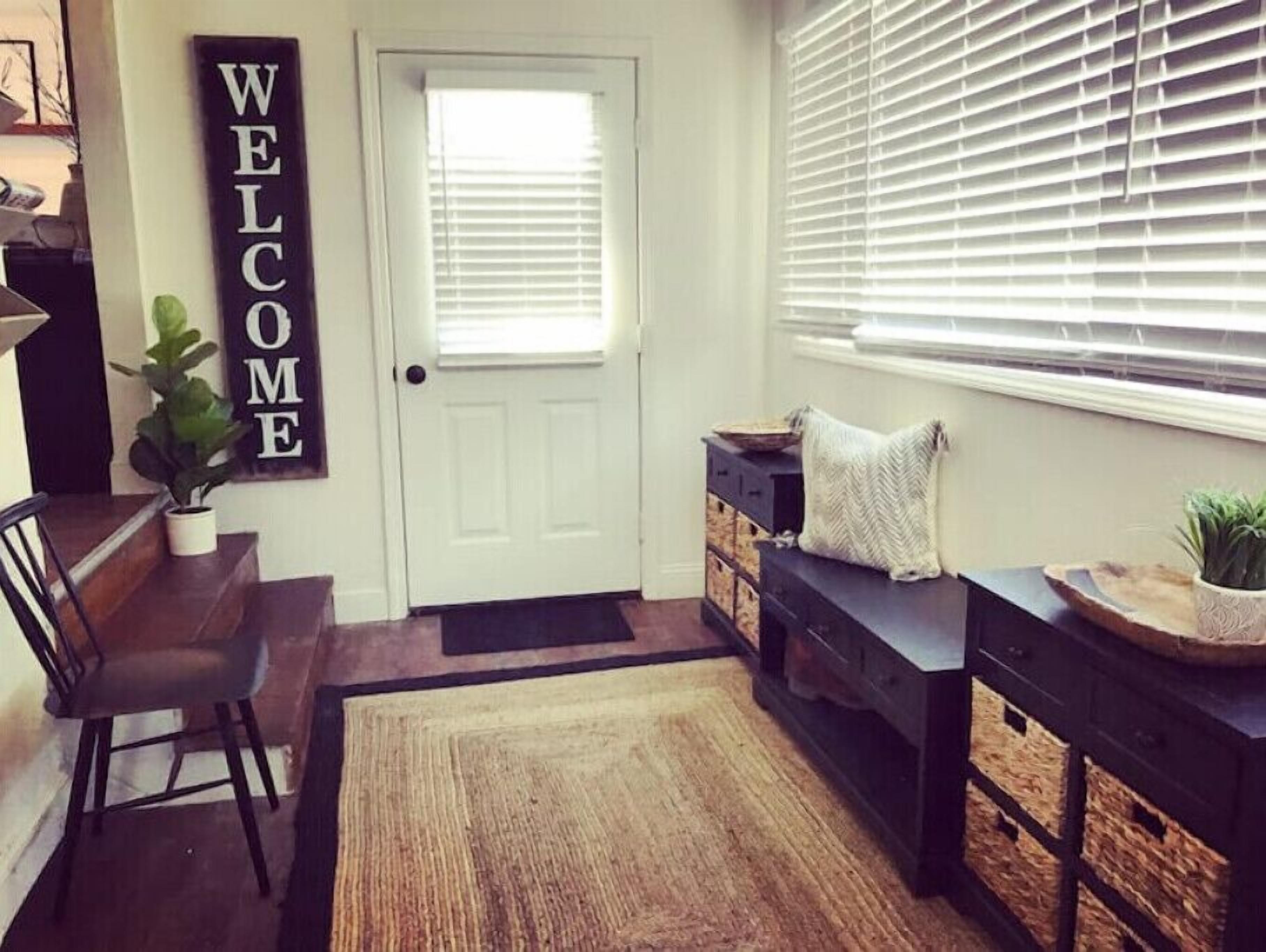 A well-lit entryway with a white door, a 'WELCOME' sign, a bench with cushions, and a potted plant.