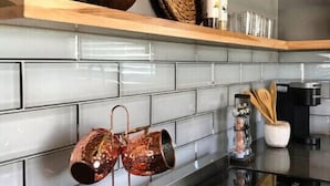 Una cocina con encimera negra, tazas de cobre colgadas en un perchero, una cesta tejida y una botella en un estante de madera.