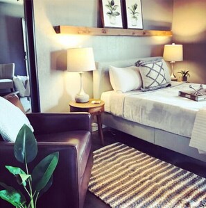 Une chambre d’hôtel avec un lit, deux lampes de chevet, une petite table, une chaise et une plante.