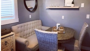 Un salón con una mesa redonda, dos sillas de mimbre, un sofá capitonado, un espejo y estantes con copas de vino y adornos.