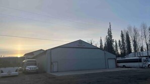 A large industrial building with a flat roof and a single garage door, surrounded by parked vehicles and trees. - Chena marina guest apartment.  (Fairbanks)