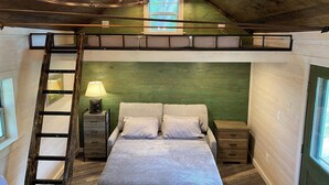 A loft-style bedroom with a loft bed, a ladder, a bedside table with a lamp, and a view of the outdoors through a window.
