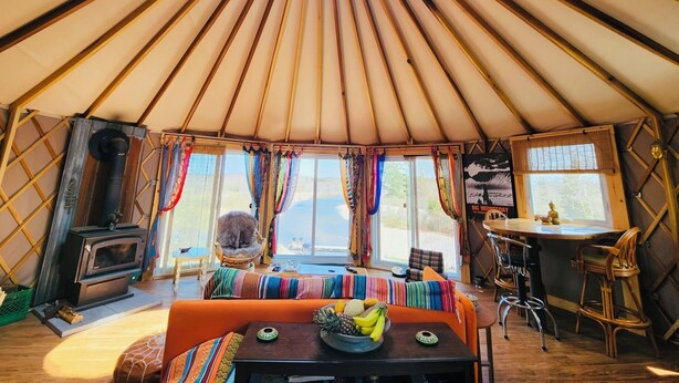 Interior de una yurta con una zona de estar central, una pequeña estufa de cocina y una mesa de comedor con fruta.