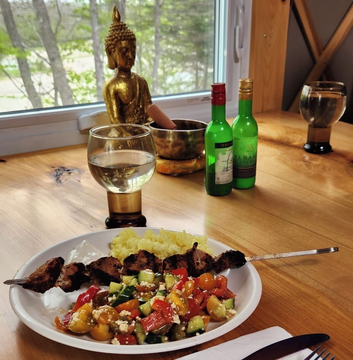 A dining table with a Buddha statue, a bowl, a plate of food, and two bottles of wine.