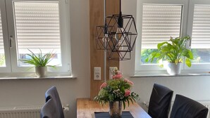 Uma sala de jantar com mesa de madeira, cadeiras pretas, luminária pendente e plantas em vasos junto às janelas.