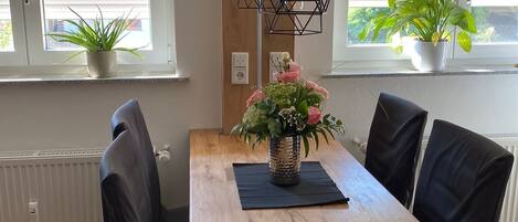 A dining area with a wooden table, black chairs, a hanging light fixture, and potted plants by the windows.