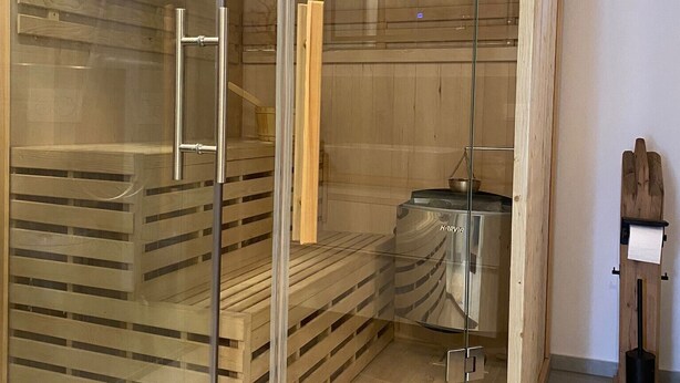 Uma sauna com divisória de vidro, bancos de madeira e um espaço visível com aquecedor e relógio.