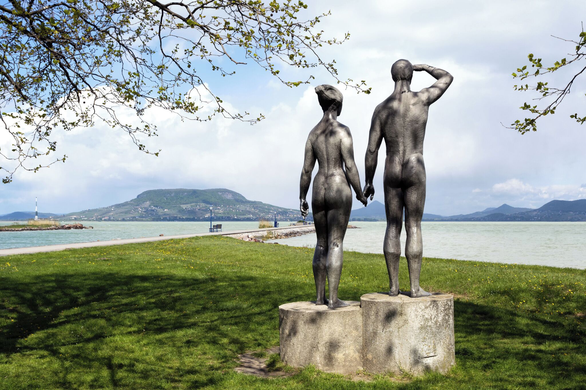 Een standbeeld van twee figuren op een platform aan een meer, met een berg en vuurtoren in de verte.