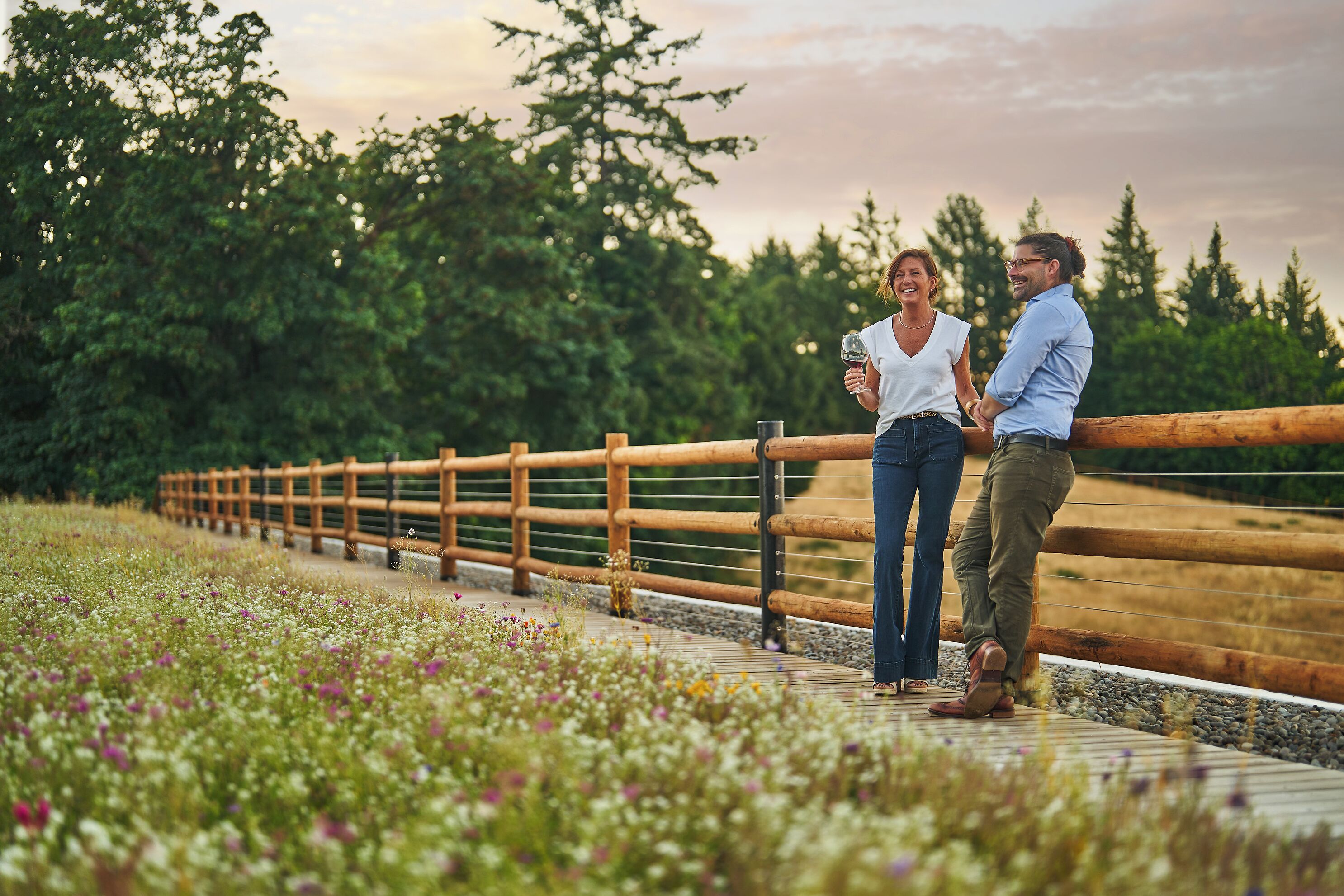 Two people standing on a wooden deck with a wooden railing, surrounded by greenery and flowers.