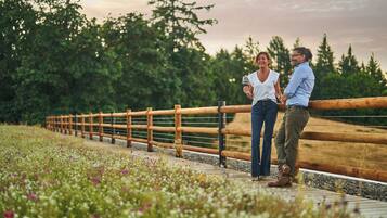 Two people standing on a wooden deck with a wooden railing, surrounded by greenery and flowers.