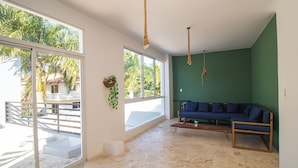 A spacious room with a stone floor, a blue sofa, and large windows offering a view of palm trees and a white building. - Villa Boho Chic (Juan Dolio)