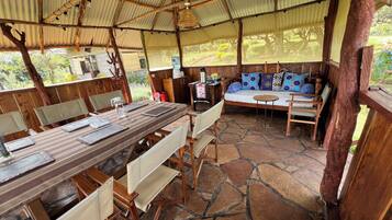 A rustic outdoor dining area with a long wooden table, cushioned chairs, and a stone-tiled floor.