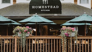 Un edificio con un letrero que dice 'Homestead Kitchen' sobre un área de asientos al aire libre techada, con sombrillas y macetas de flores.