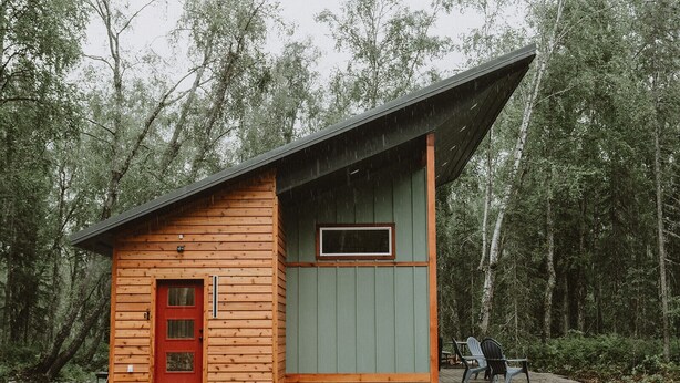 Una cabaña pequeña con puerta roja y ventana, rodeada de un bosque.