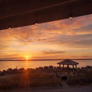 A sunset view from a covered area with a wooden gazebo and a sandy beach.