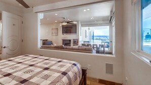 A bedroom with a large window, a bed with a checkered bedspread, and a view of a balcony with outdoor seating.
