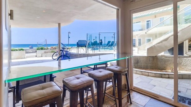 A modern bar area with a glass table and leather stools, overlooking a beach and a staircase leading to an upper level.