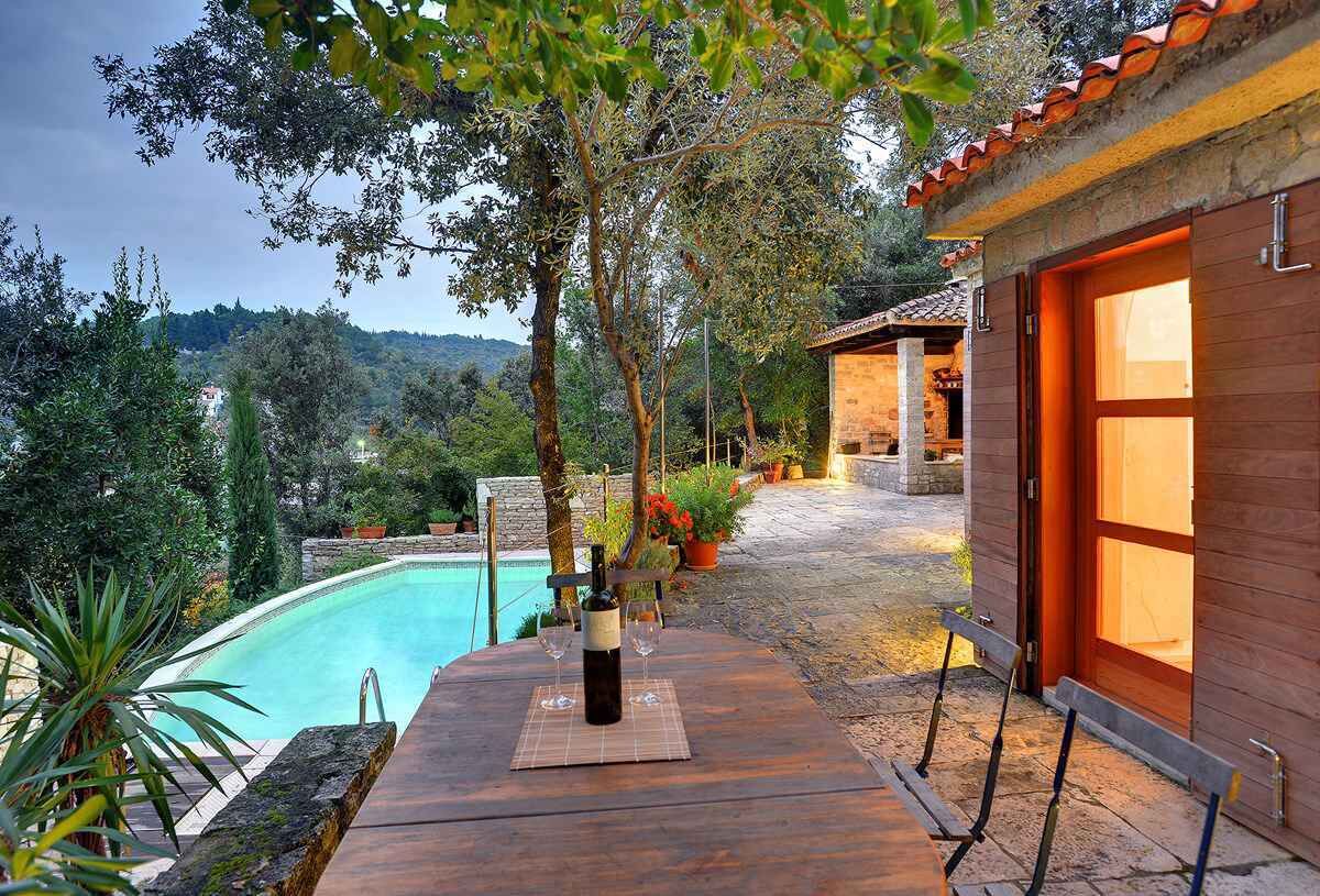 Una terraza de madera con piscina, rodeada de árboles y un sendero de piedra que lleva a un edificio con grandes ventanales.