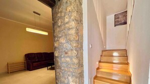 Eine Treppe mit Holzbodenstufen führt zu einer oberen Etage, eine Steinsäule, ein dunkelbraunes Ledersofa und ein heller Fliesenboden sind zu sehen.