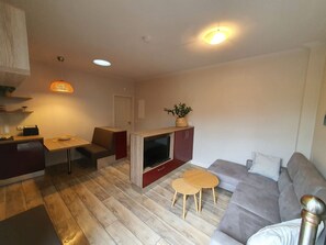 Ein modernes Wohnzimmer mit grauem Sofa, einem kleinen Holztisch, einem Fernseher und einer Küchenzeile mit Holzschrank und einer Pendelleuchte.