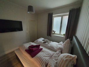 A bedroom with two beds, a TV mounted on the wall, a window with curtains, and a hanging light fixture. - Altstadtdomizil Wohnung Helin - Pure Entspannung in der Altstadt Borkums! - Alte Schulstraße (Borkum)