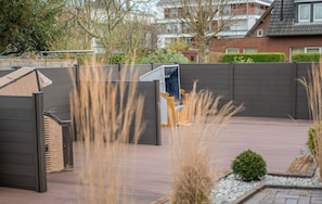A modern outdoor patio with a wooden bench, a metal gate, and a view of residential buildings in the background. - Altstadtdomizil Wohnung Helin - Pure Entspannung in der Altstadt Borkums! - Alte Schulstraße (Borkum)