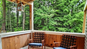 Una terraza de madera con dos mecedoras y una mesita, con vista a un bosque frondoso.