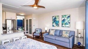 A hotel room with a bed, a sofa, a chair, a small table, a mirror, artwork on the wall, and a ceiling fan. - Endless & Elegant Vistas (Kihei)