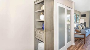 A hallway with a built-in storage unit, a door leading to a living room, and a grey sofa. - Endless & Elegant Vistas (Kihei)