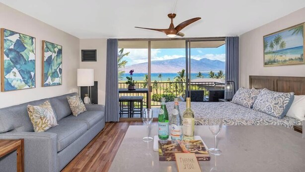 A hotel room with a balcony, a sofa, a bed, and a table with drinks and a thank you card. - Endless & Elegant Vistas (Kihei)