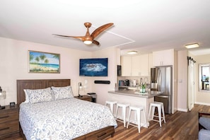 Ein Schlafzimmer mit Bett, Deckenventilator, einem Strandbild und einer Küchenzeile mit Kühlschrank und Barhocker.