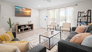 A modern living room with a flat-screen TV, a ceiling fan, a bookshelf, and a large window with curtains. - PuttPutt, Outdoor TVs & BBQ Bar, Pool & Spa! 5 mins from Old Town! (Scottsdale)