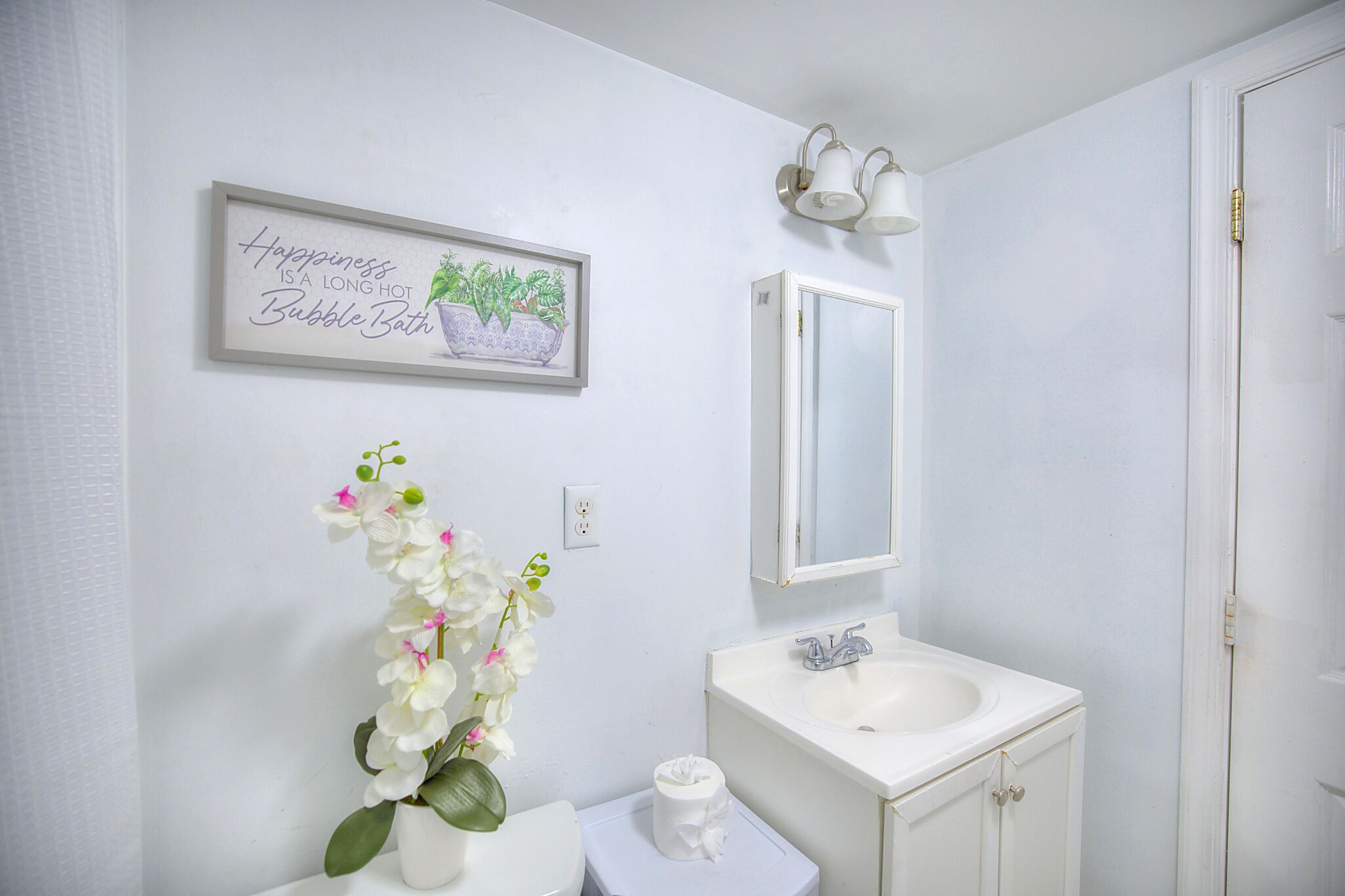 A bathroom with a white sink, mirror, and toilet. There is a framed picture on the wall and a potted plant.