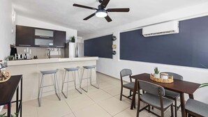 Una cuina moderna amb una illa central, tamborets de bar, una taula de menjador amb caires, un ventilador de sostre i un aparell d'aire condicionat.